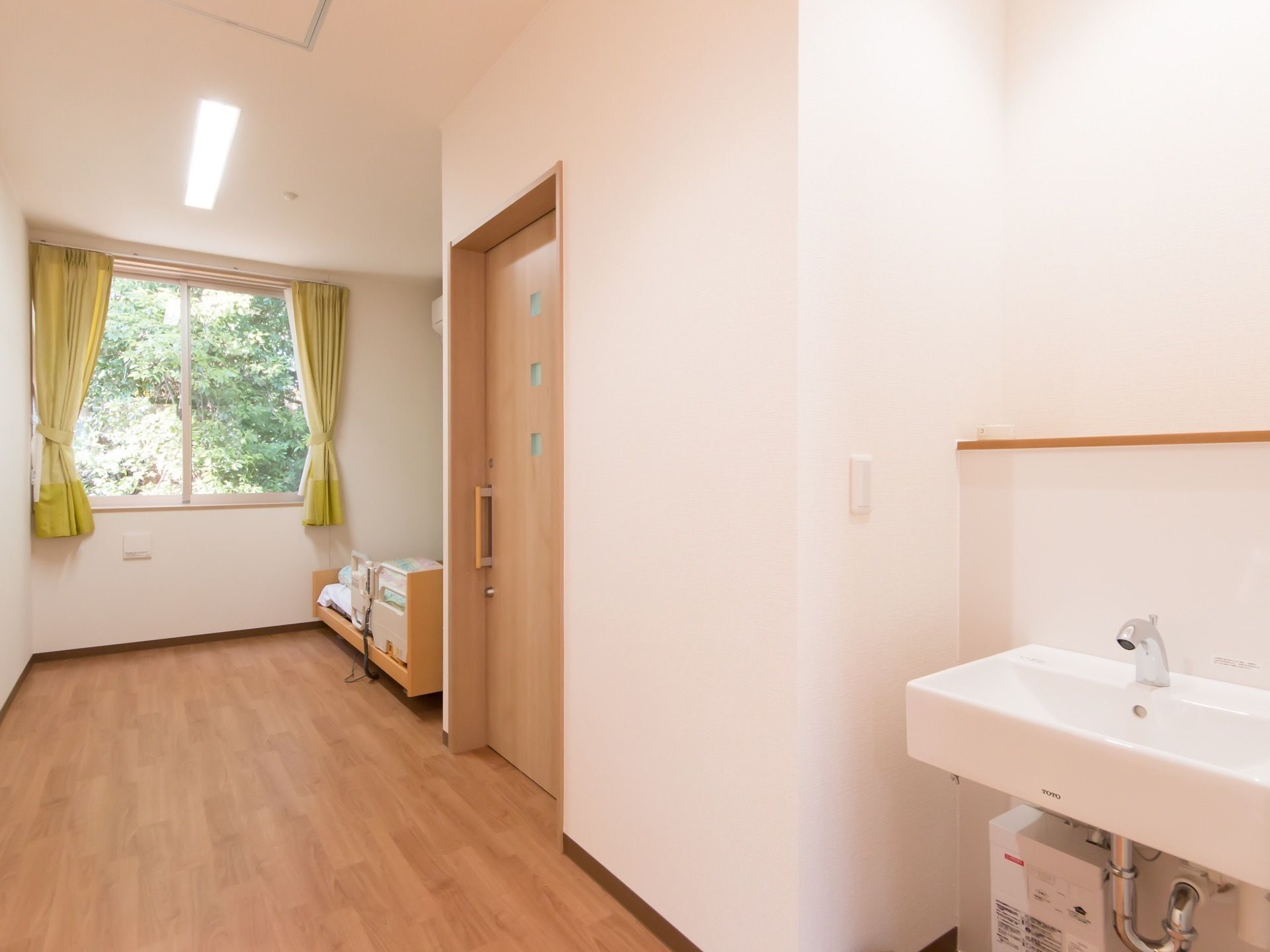 居室はナチュラルな木目が基調となっている。手前には手洗い場がある。奥には窓が設けられており、その下にベッドが置かれているのが見える。
