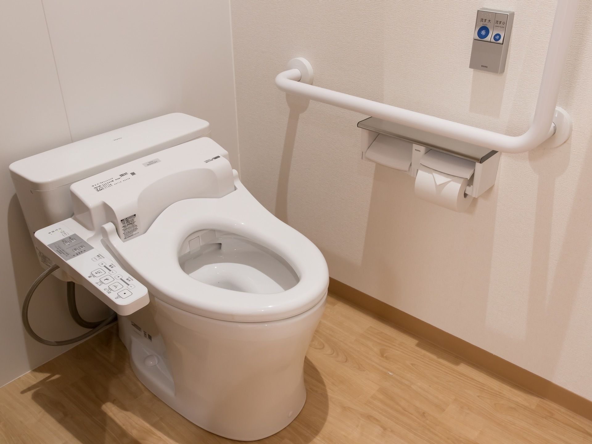 トイレは温水洗浄便座完備で、左手の壁に操作パネルと大きな手すり、トイレットペーパーホルダーが設置されている。
