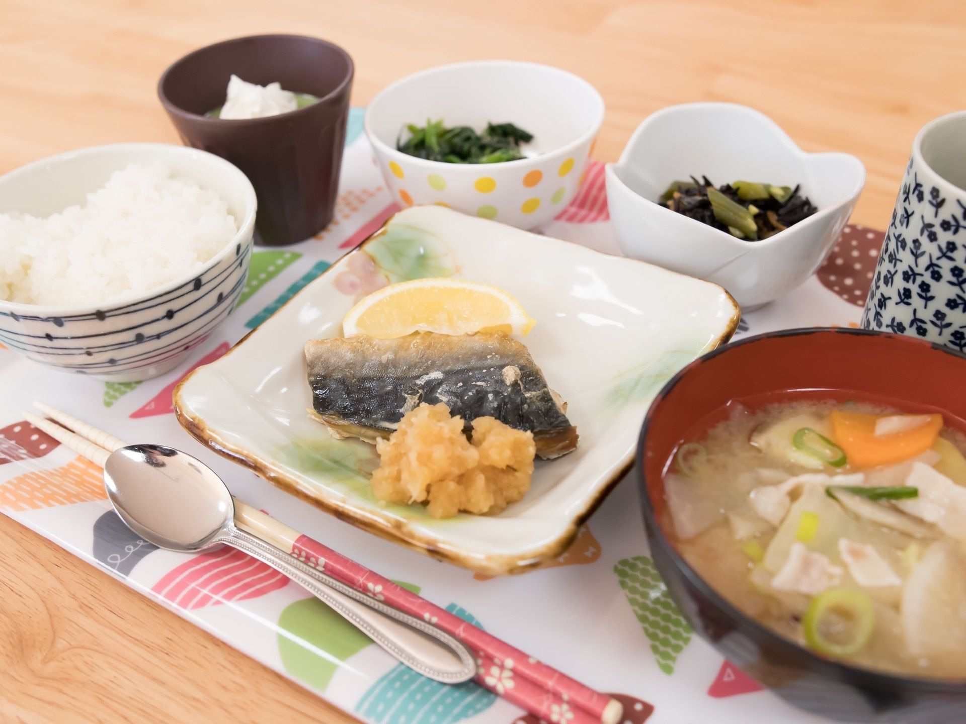 白地にカラフルな模様が描かれたプレートに、茶碗や小鉢が載っている。手前に箸とスプーンが置かれている。