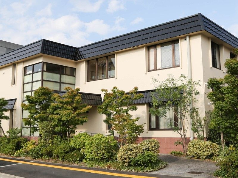 グレーの屋根が付いた2階建ての建物はベージュの外壁で、敷地内にはたくさんの緑が植えられている。