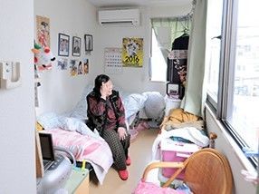居室のベッドに座っている女性入居者。エアコンやいす、写真などが置かれている。