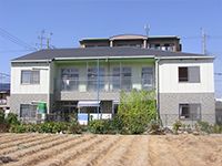 広い畑がホームの前に広がっている。2階建ての建物は白とグレーでダークグレーの屋根が付いている。