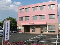広い駐車場があり、日当たりのいい場所に立っているピンクの外壁の3階建ての建物。敷地内には木や花も植えられている。