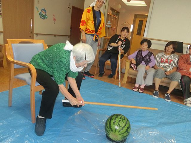 白髪の高齢女性がいすに座ったままスイカ割りに挑戦している。目隠しはしておらず、首の周りにタオルがある。
