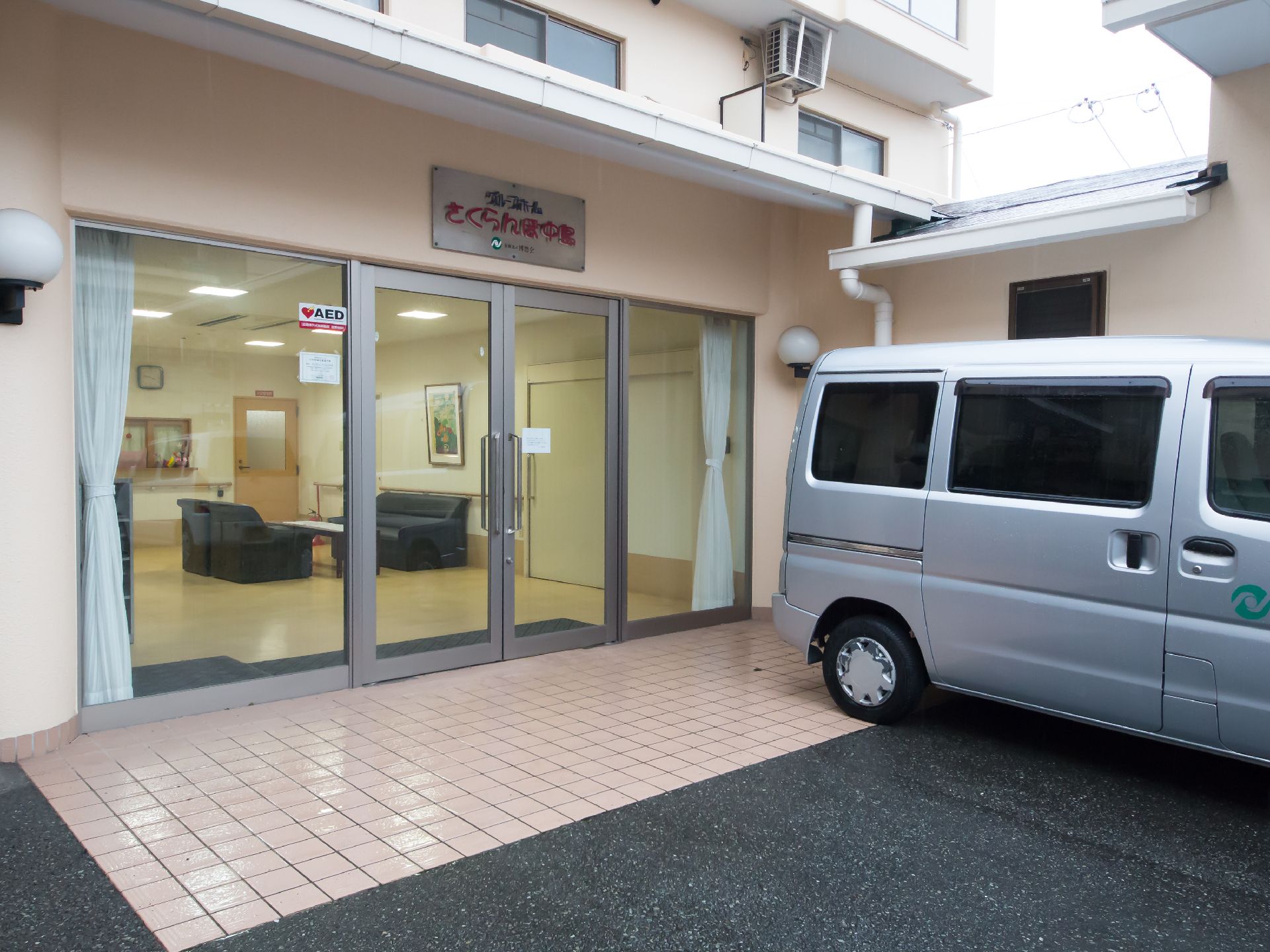 両開きのガラス扉の入口である。入口前にシルバーの軽乗用車が停まっている。玄関の上部の壁に施設名が表示されている。