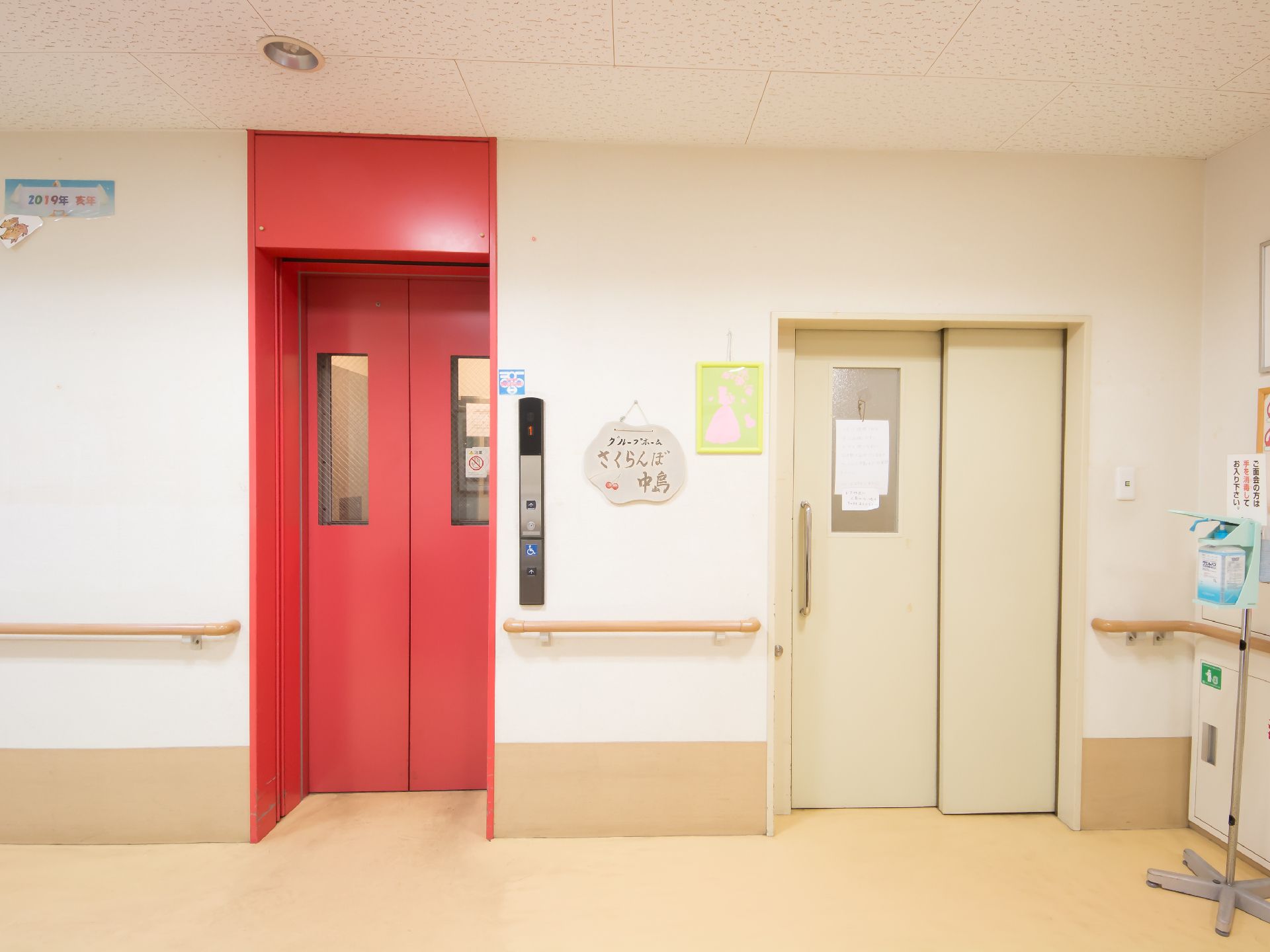 奥の壁に赤い扉のエレベーターがあり、その右側に引き戸扉がある。２枚の扉の間の壁には絵画が飾られている。