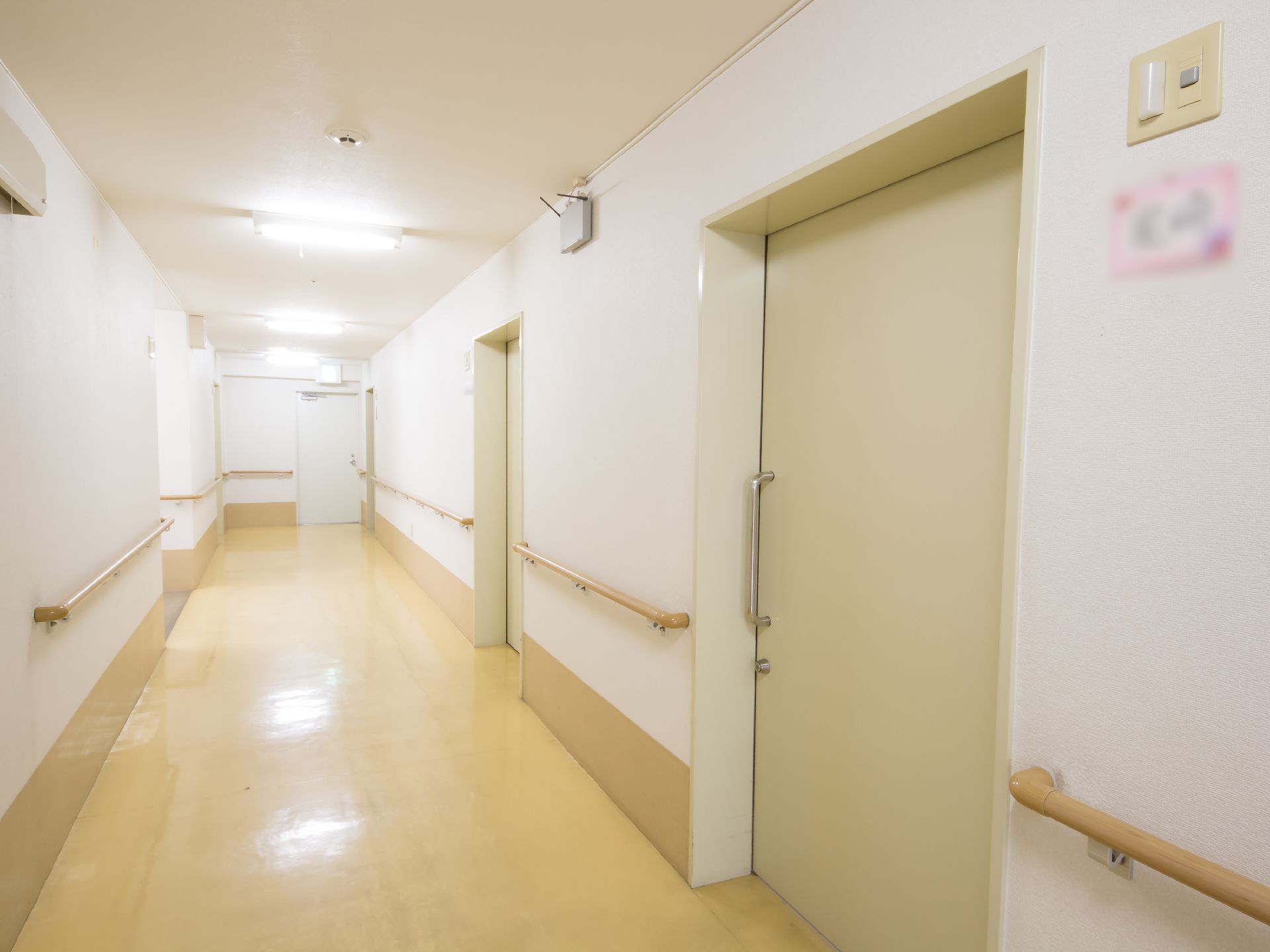 白い壁の廊下で右側にクリーム色のドアが並んでいる。引き戸のドアの横の壁にネームプレートが貼られている。