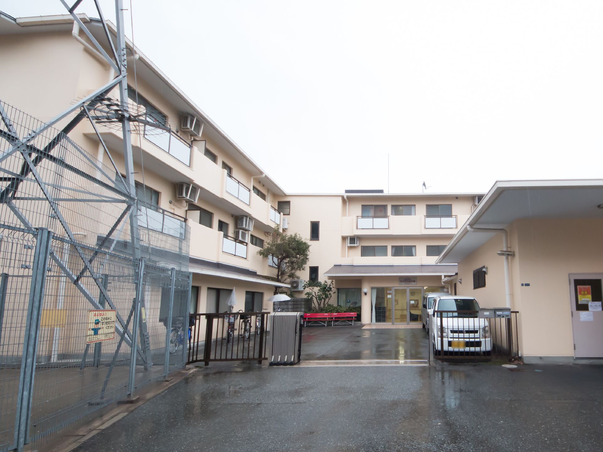 薄いベージュの３階建ての建物で、１階部分は別の建物が右手にせり出している。入口に黒い鉄製の門がある。