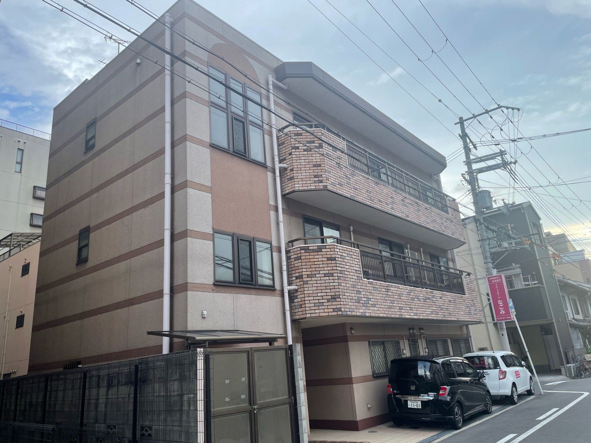 施設の建物は３階建てで、２階と３階に同じ素材同じ形状のベランダが付いている。一見低層マンション風の外観で、建物の端に看板が設置されている。