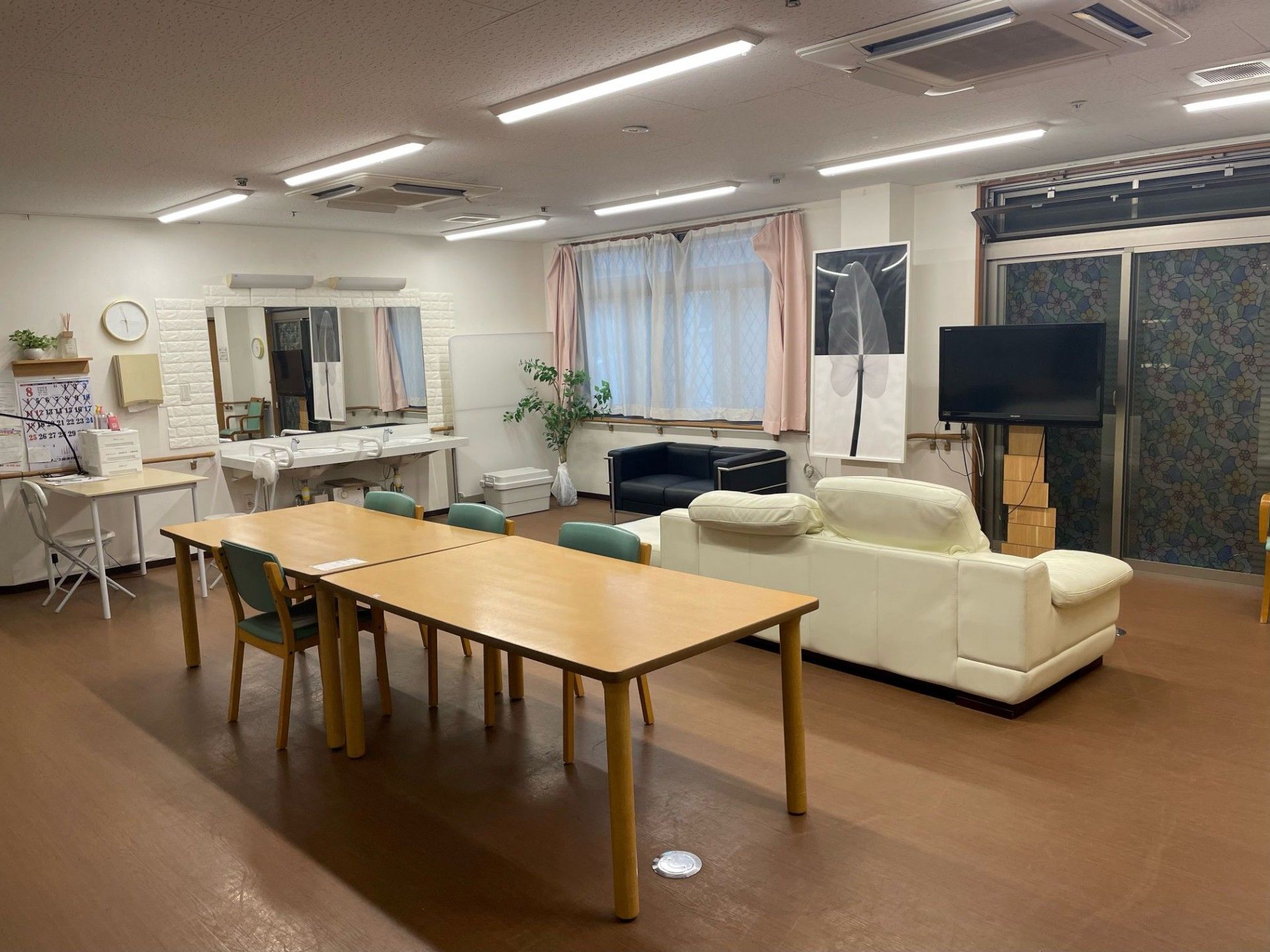 広い共有ペースにはテーブルと椅子が配置され、正面には窓と窓の間の壁際にテレビが置いてある。天井には多数の照明が設置され部屋を照らしている。