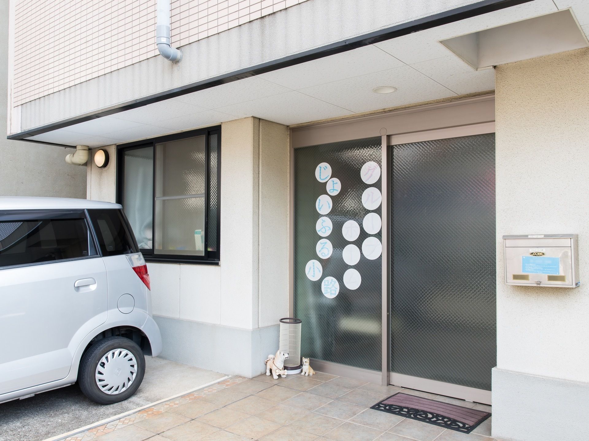 金網の入ったガラス扉のある玄関。地面はタイル張りで、玄関マットが敷かれている。ガラスドアには丸い紙に施設名か書かれたものが貼られている。