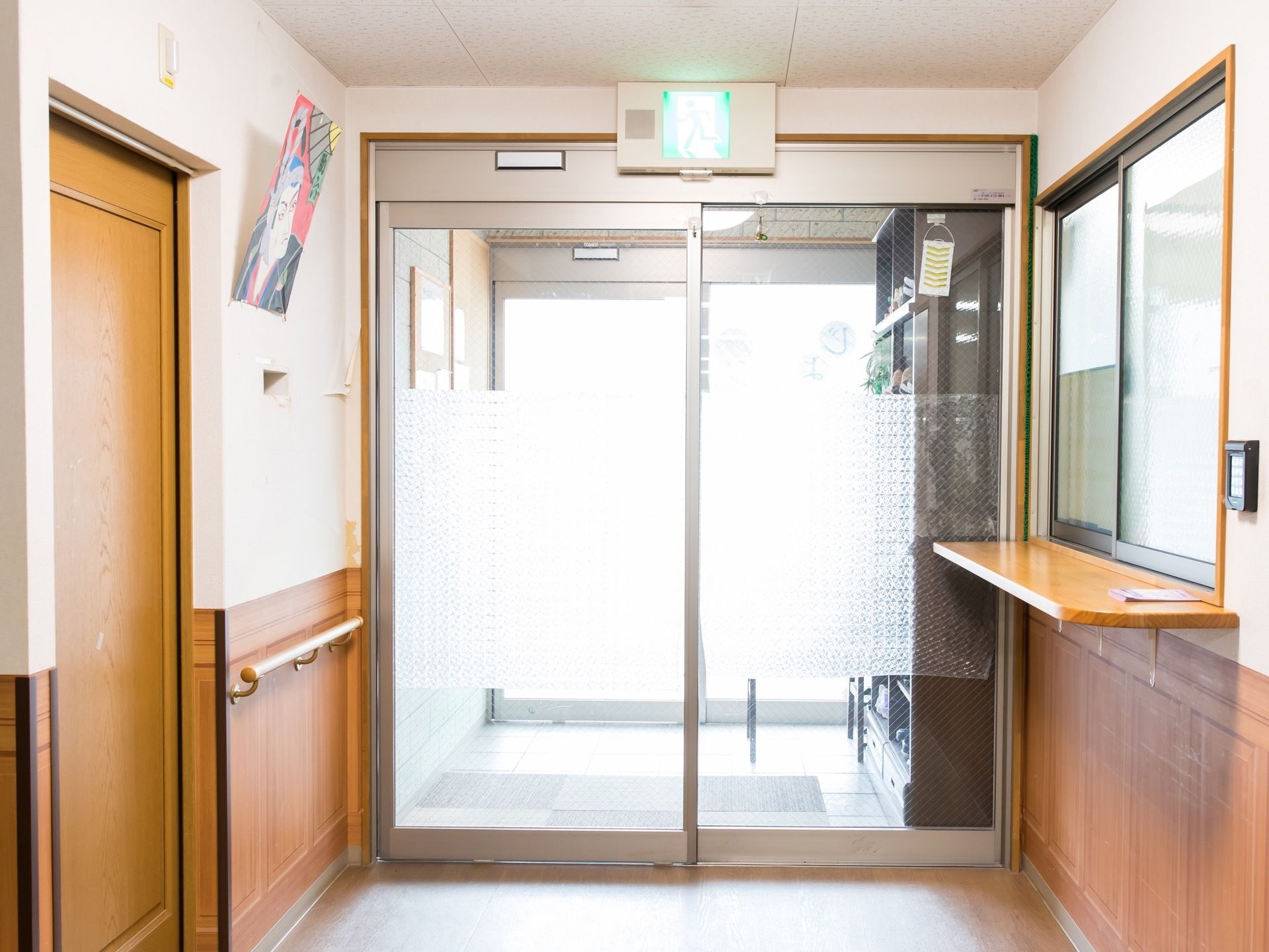 ガラス張りの２重扉になった玄関がある。大きな下駄箱が置かれ、受付窓口がある。壁には手すりが設置され、絵が飾られている。