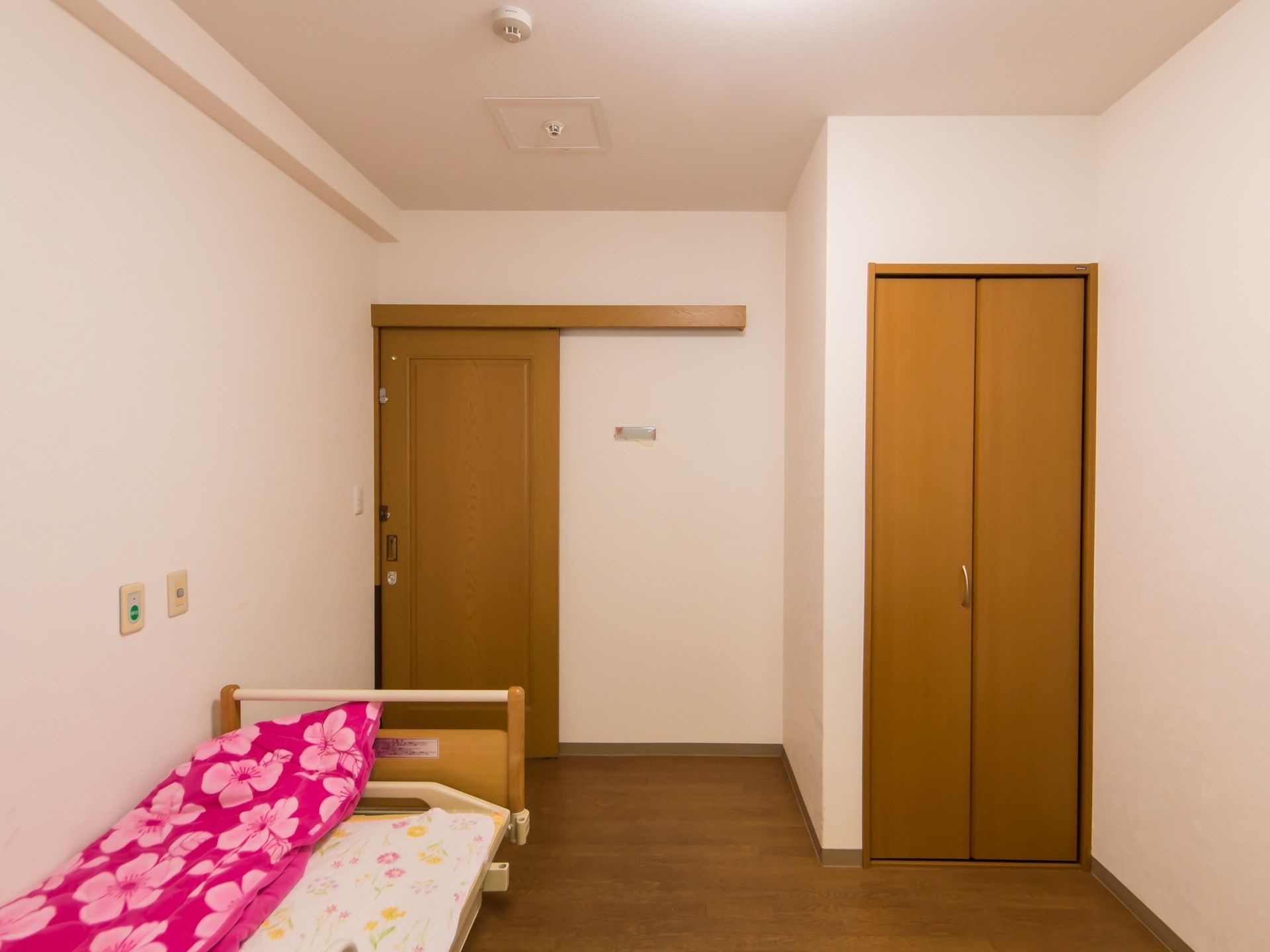 白い壁の居室がある。ライトブラウンの床で、同系色の部屋のドアとクローゼットがある。壁際には介護用ベッドがある。