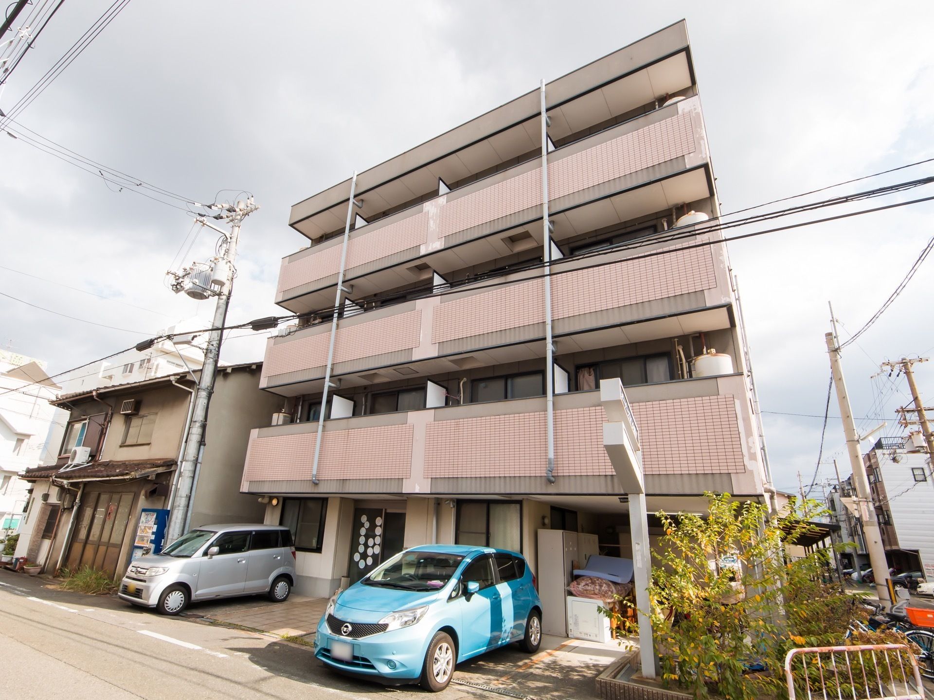ベージュのタイルの壁の４階建ての建物がある。角地に建っており、隅に植え込みがある。手前は駐車場になっている。