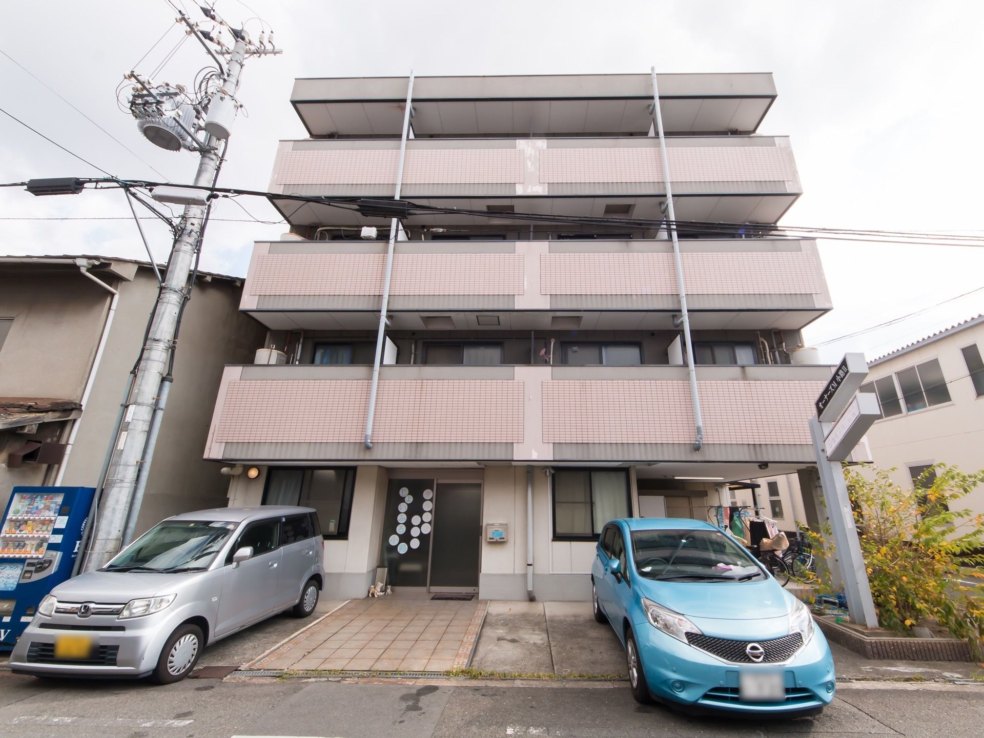ベージュのタイルの壁の４階建ての建物がある。玄関へと続く通路はタイル張りのスロープになっている。正面には車が数台停まっている。