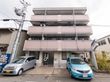 ベージュのタイルの壁の４階建ての建物がある。玄関へと続く通路はタイル張りのスロープになっている。正面には車が数台停まっている。