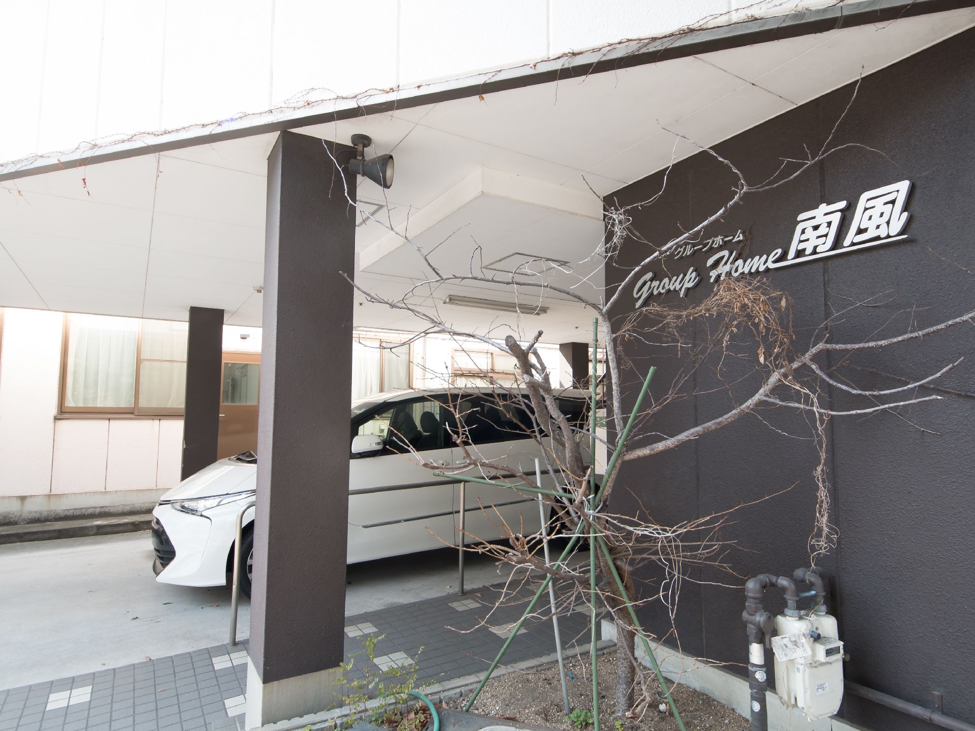 黒い壁の玄関に白い文字で施設名が表示されている。入口前にあるスロープの向こうに白い乗用車が１台停められている。