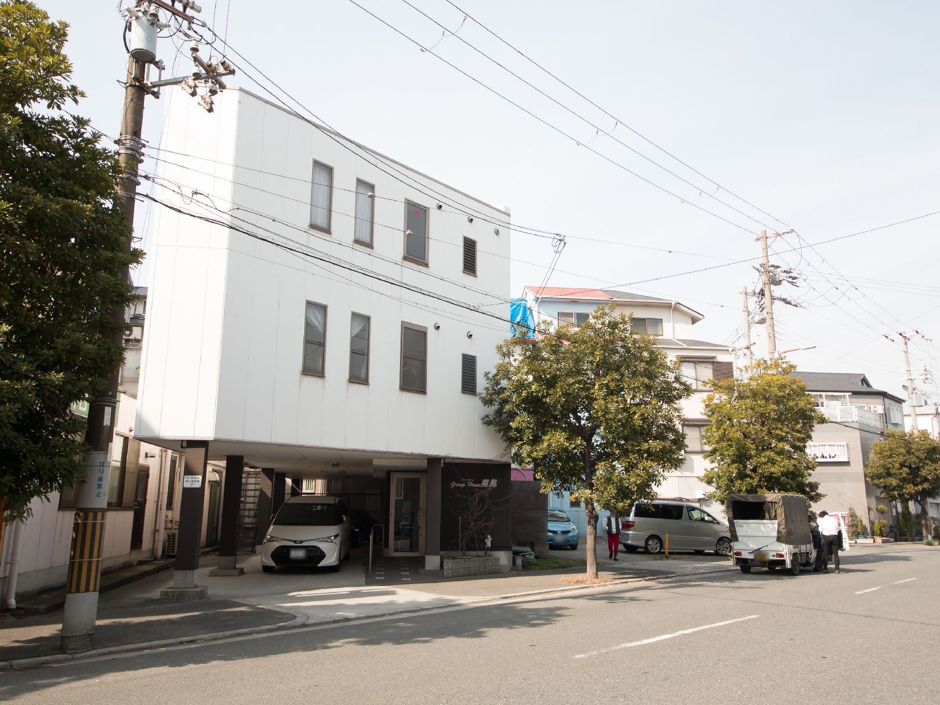大通りに面した白い外壁の建物で、３階建てである。施設前には樹木が植えられている。１階部分に自動車が停まっている。