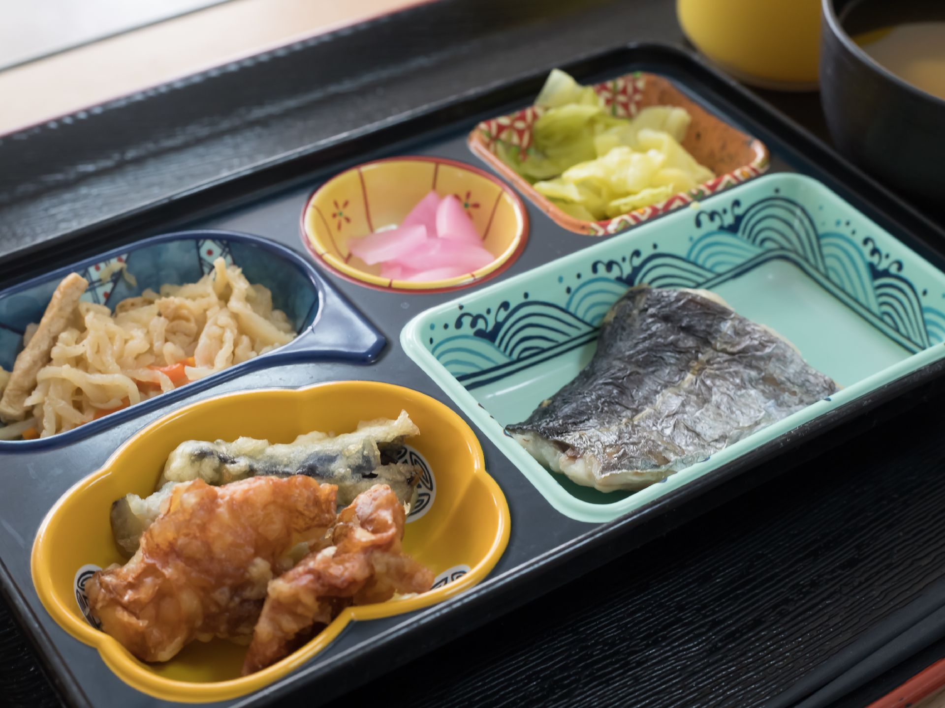 黒いトレイにさまざまな和柄の器が並んでいる。焼き魚、揚げ物と切り干し大根、お浸しや漬物が盛られている。
