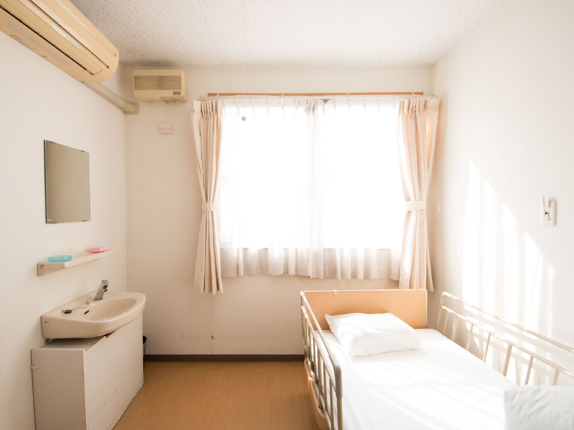 居室の奥に大きな窓があり、カーテンがかかっている。窓の前に落下防止柵のついた介護用ベッドが置かれている。