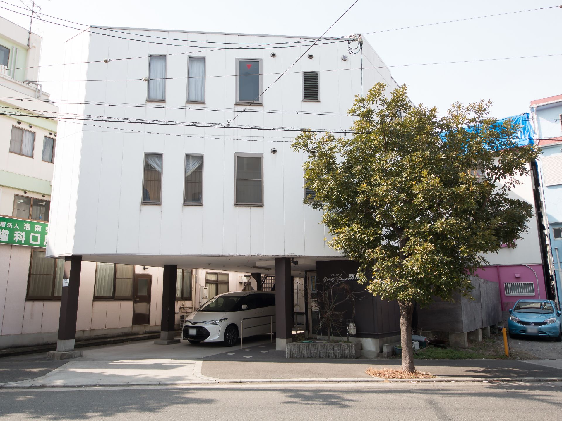 白い壁の建物で１階部分に駐車スペースのある３階建ての施設である。施設前に街路樹があり、施設の隣に歯科医院がある。