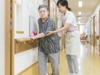 廊下を手すり伝いに歩いている高齢女性と付き添いのエプロンを付けた女性スタッフ。