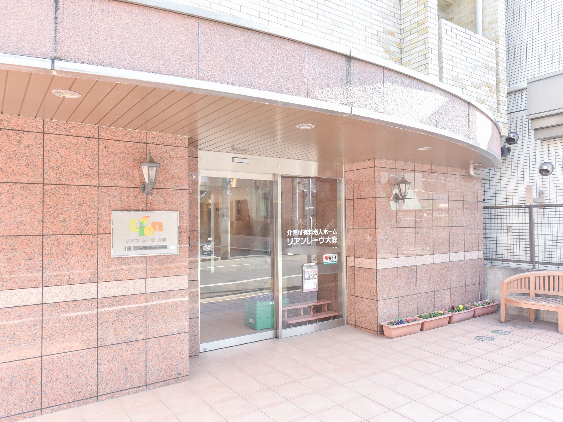 建物玄関はガラスの自動ドアで施設名が表示してある。ドアの両サイドに玄関灯がある。庇部にも埋込式照明がある。玄関回りの外壁はピンク系である。