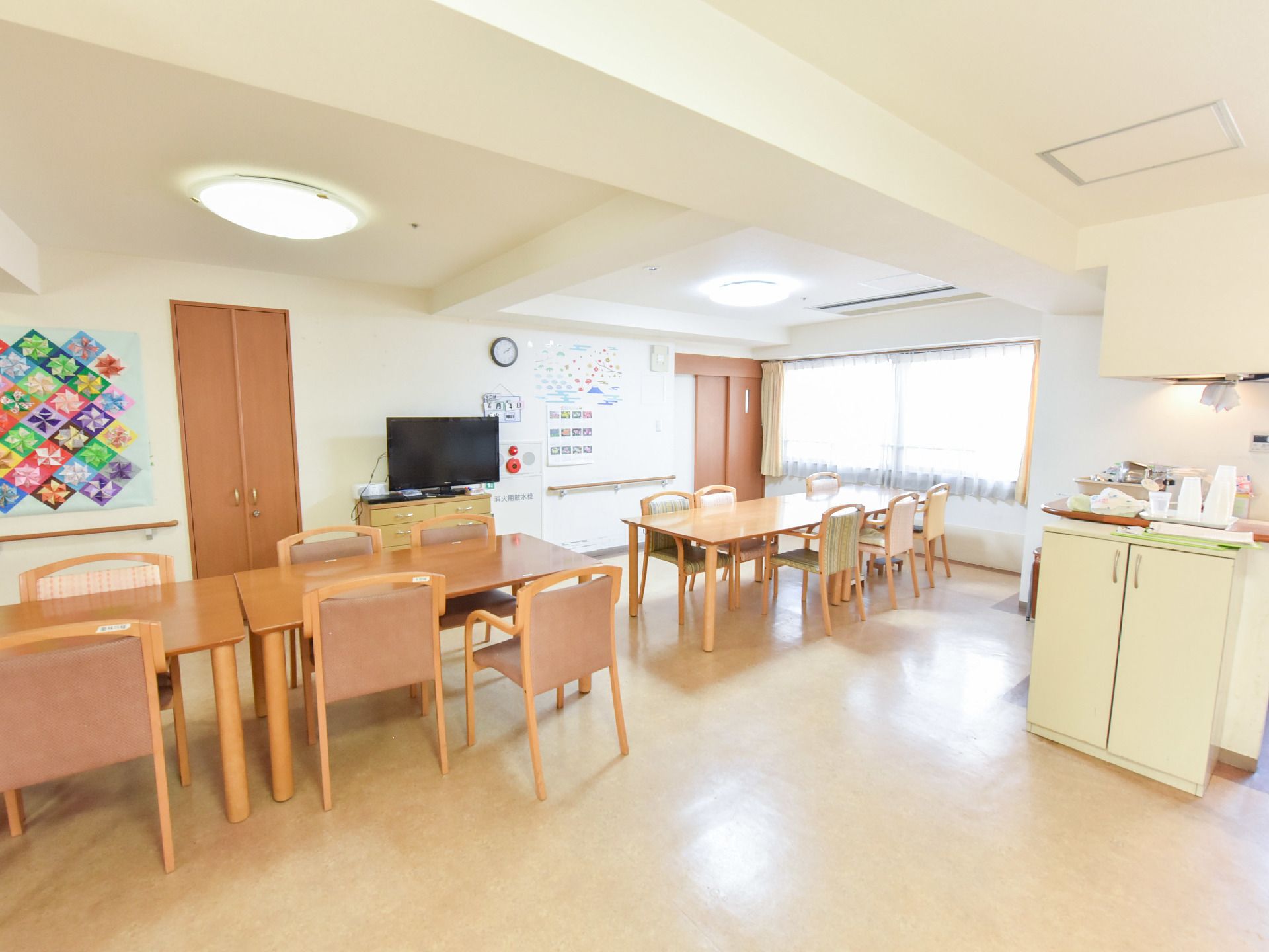 白と茶系でまとめられた内装と一面窓のリビング。長テーブルと椅子が並べられている。中央には、テレビが置いてある。