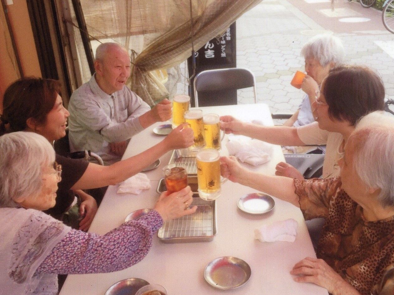 路面にある飲食店で、入居者様とスタッフがテーブルを囲んでいる。丁度ビールジョッキで乾杯している最中で、楽しい様子が伝わってくる。