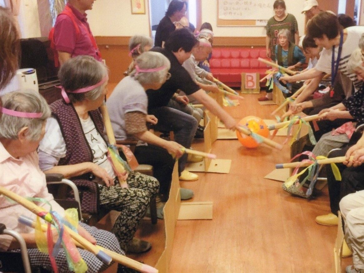入居者様が頭に鉢巻をして手には棒を持ち、向かい合わせで座っている。ビーチボールを棒で転がすゲームを行っており、熱が入り夢中な様子である。