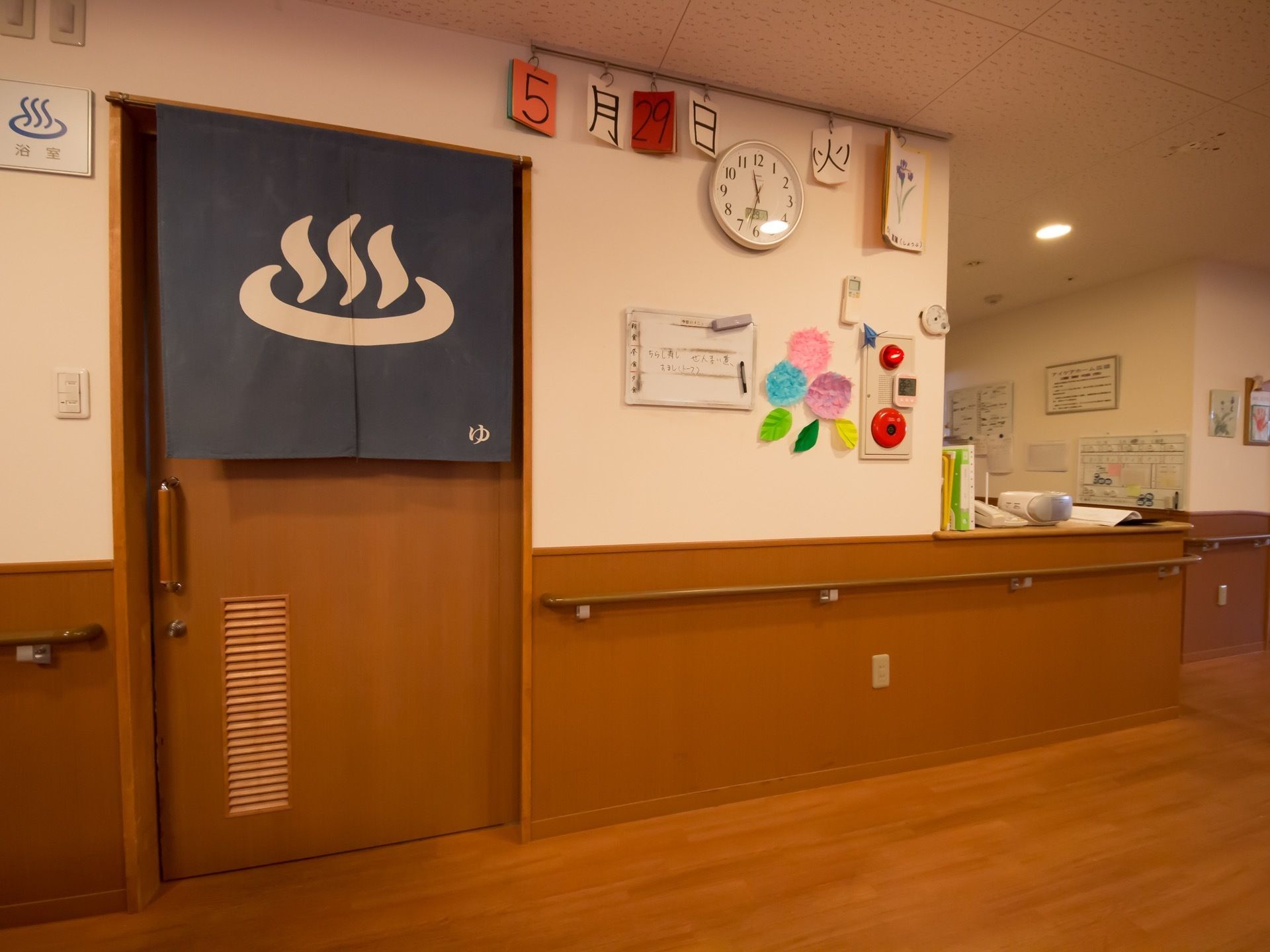 浴室前のドアには銭湯のマークが描かれたのれんが掲げられている。壁側には手すりが設けてあり、壁には時計や防災ベルのボタンが設置してある。