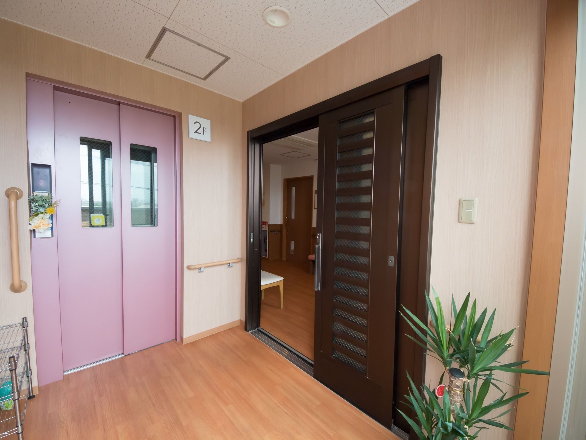 エレベーターの扉は淡いピンク色のデザインで、左側には白や黄色の花が飾ってある。両側の壁には、手すりが設置されている。
