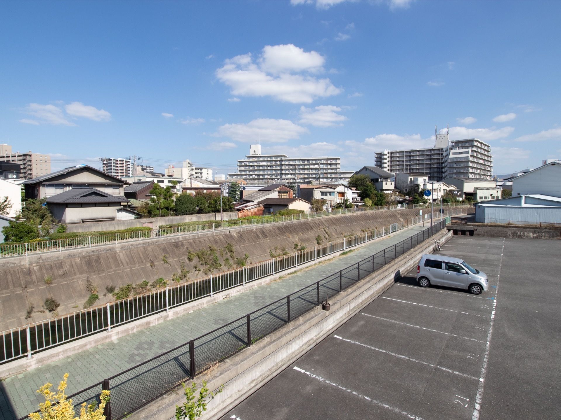 広々とした駐車場がある。周辺には川と住宅街が広がっており、離れた場所にマンションが建っている。川沿いには散歩道がある。