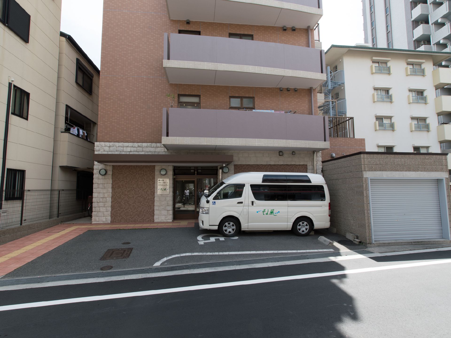 施設は茶色のタイルと白のレンガ状の外壁の4階建てのマンション風の建物。二階以上にはベランダがある。駐車場には施設の介護車両が停められている。