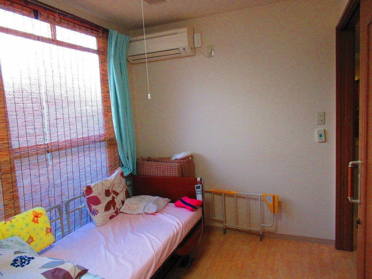 すだれが付いた大きな窓がある居室には、寝具付きのベッドが置かれ、壁にはエアコンが付いている。