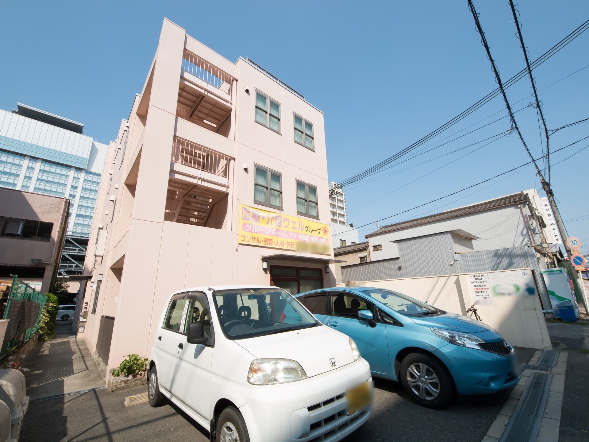 ピンクがかった色の可愛らしい外観。３階建てでシンプルな作りになっている。駐車スペースには２台の車が停まっている。