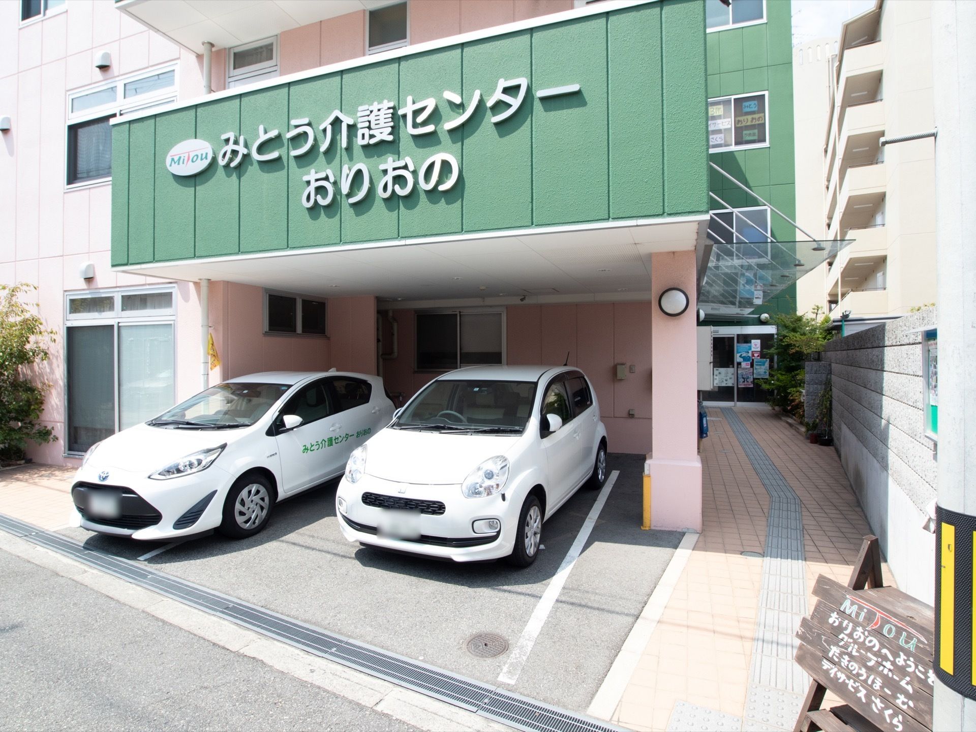 道路に面した場所に木製の案内板が用意されており、玄関までは点字ブロックが設置されている。車が2台停められている。