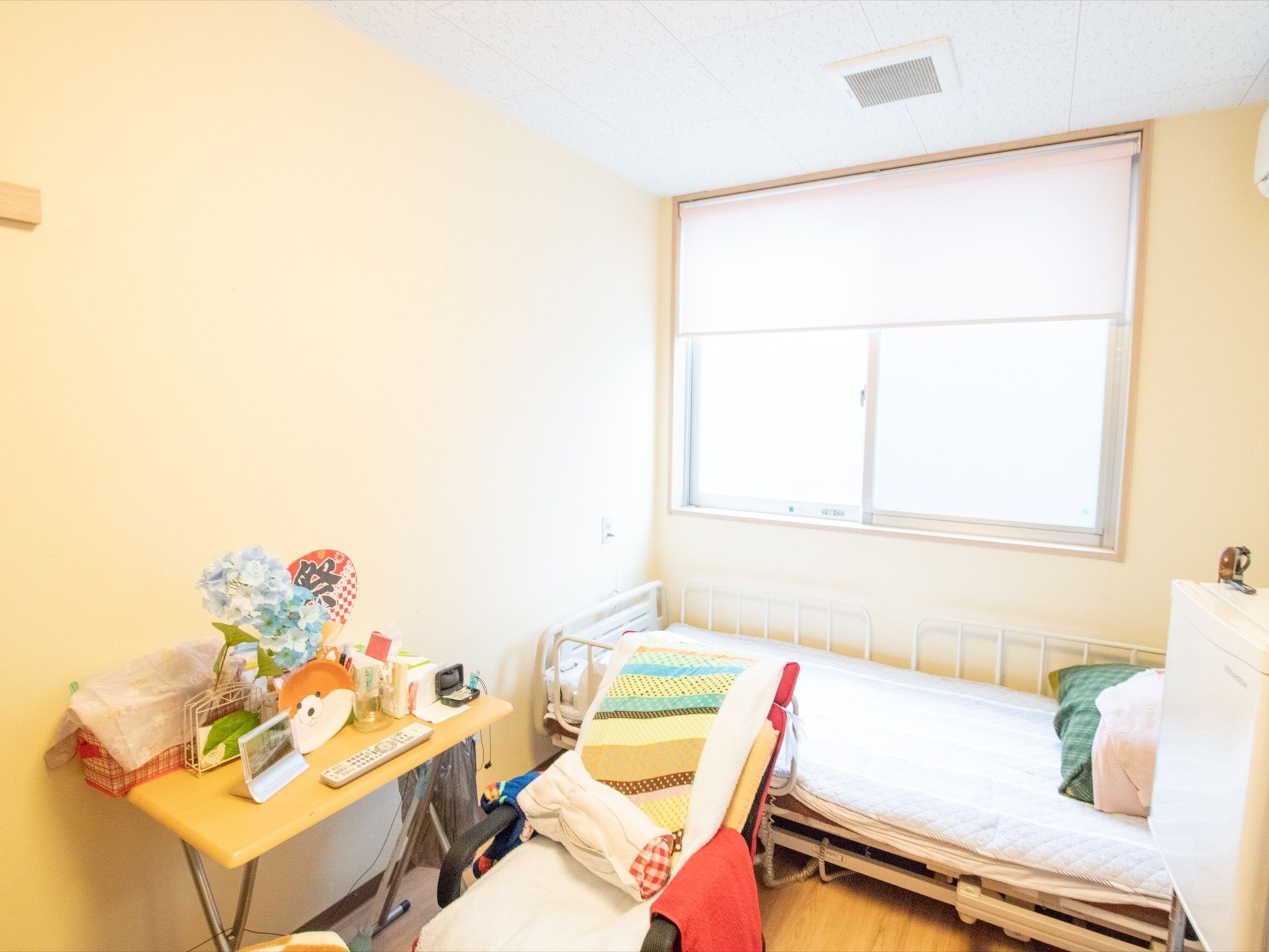窓側に電動ベッドが置かれている。窓から日差しが差し込み明るい居室になっている。テーブルの上にさまざまな小物が並んでいる。
