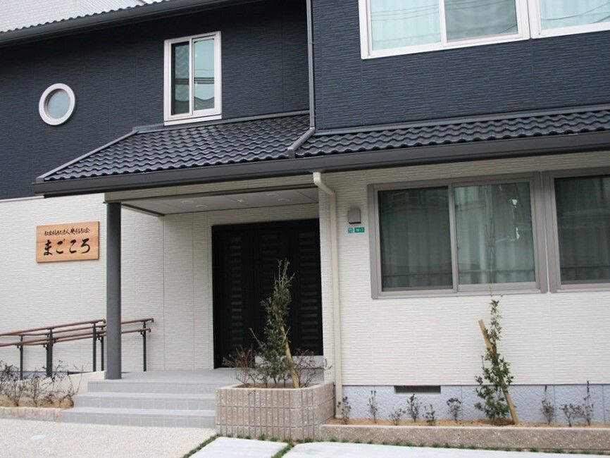 2階建ての建物はモノトーンでモダンな雰囲気。玄関には階段と手すり付きのスロープが付き、花壇もある。