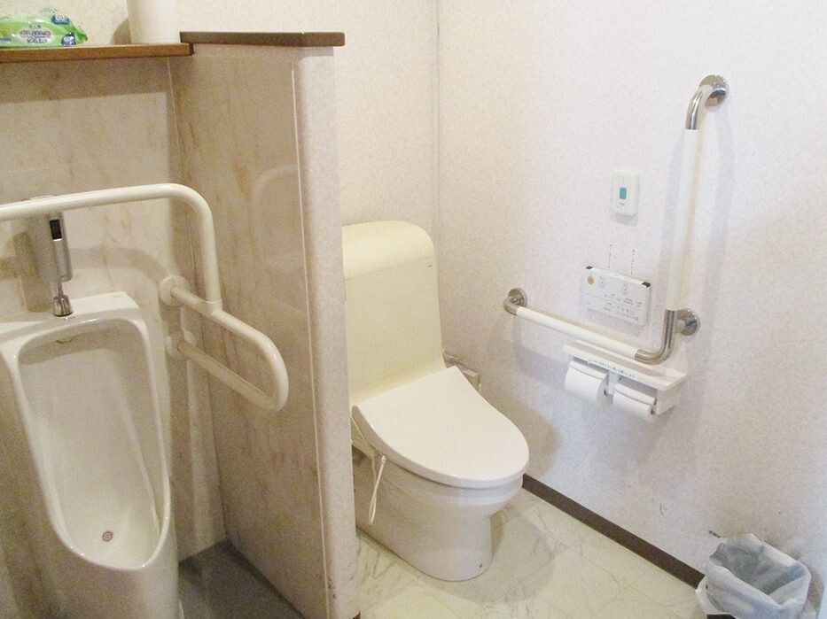 右側にはエル字型の手すりや呼び出しボタンが付いた洋式トイレがあり、壁をはさんで左側には男性用小便器がある。