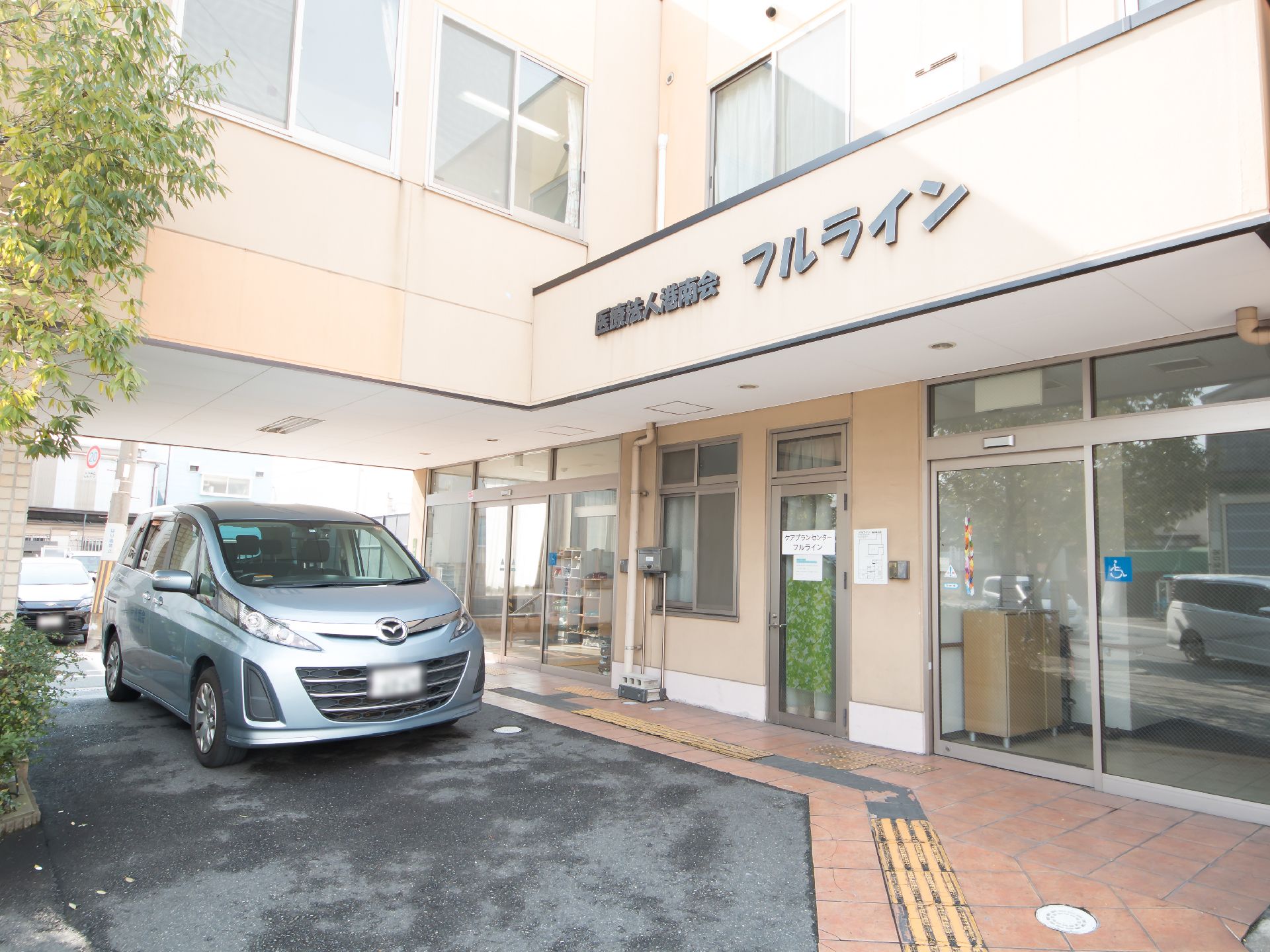 玄関前に車が停められるスペースがあり、シルバーのワゴンが停まっている。入口はガラスの自動扉になっている。