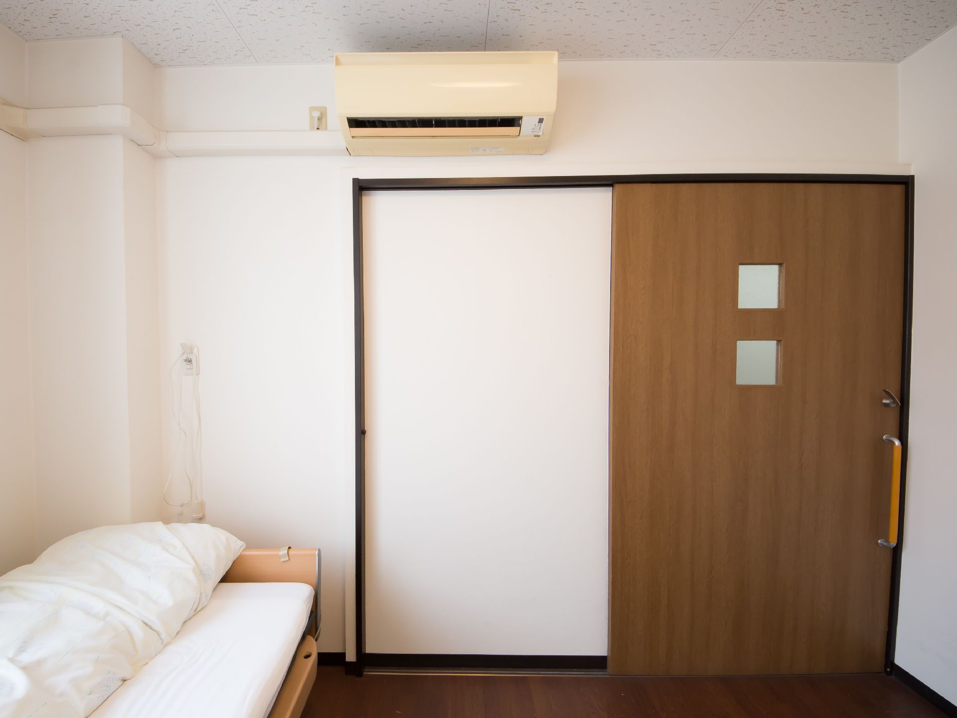 茶色の引き戸の扉があり、窓が２つ開いている。入口ドアの上にエアコンがある。壁際にベッドが置かれている。