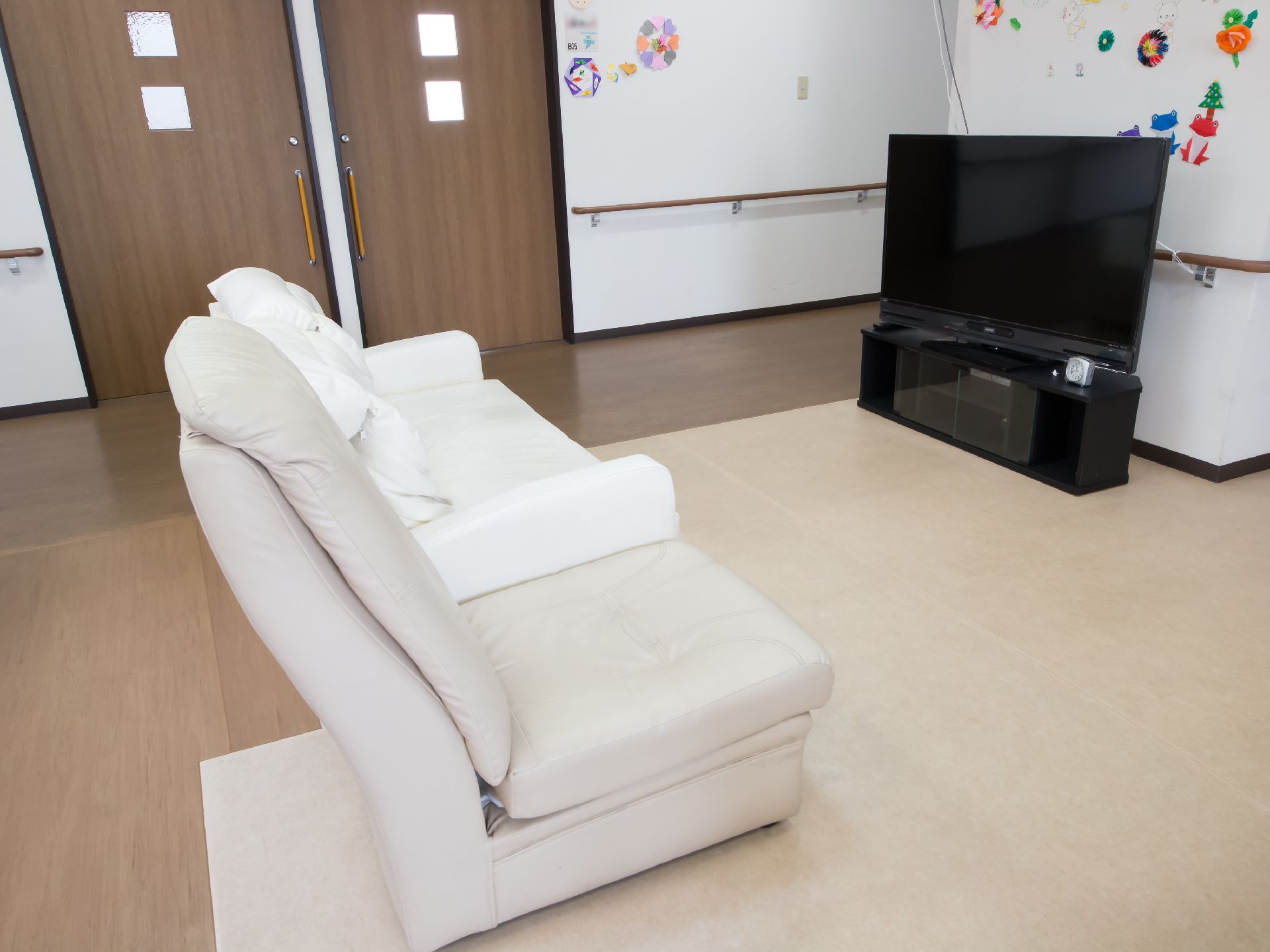 白い２人用ソファがあり、その隣に１人用のグレーのソファが並べられている。奥には居室へのドアが２か所にある。