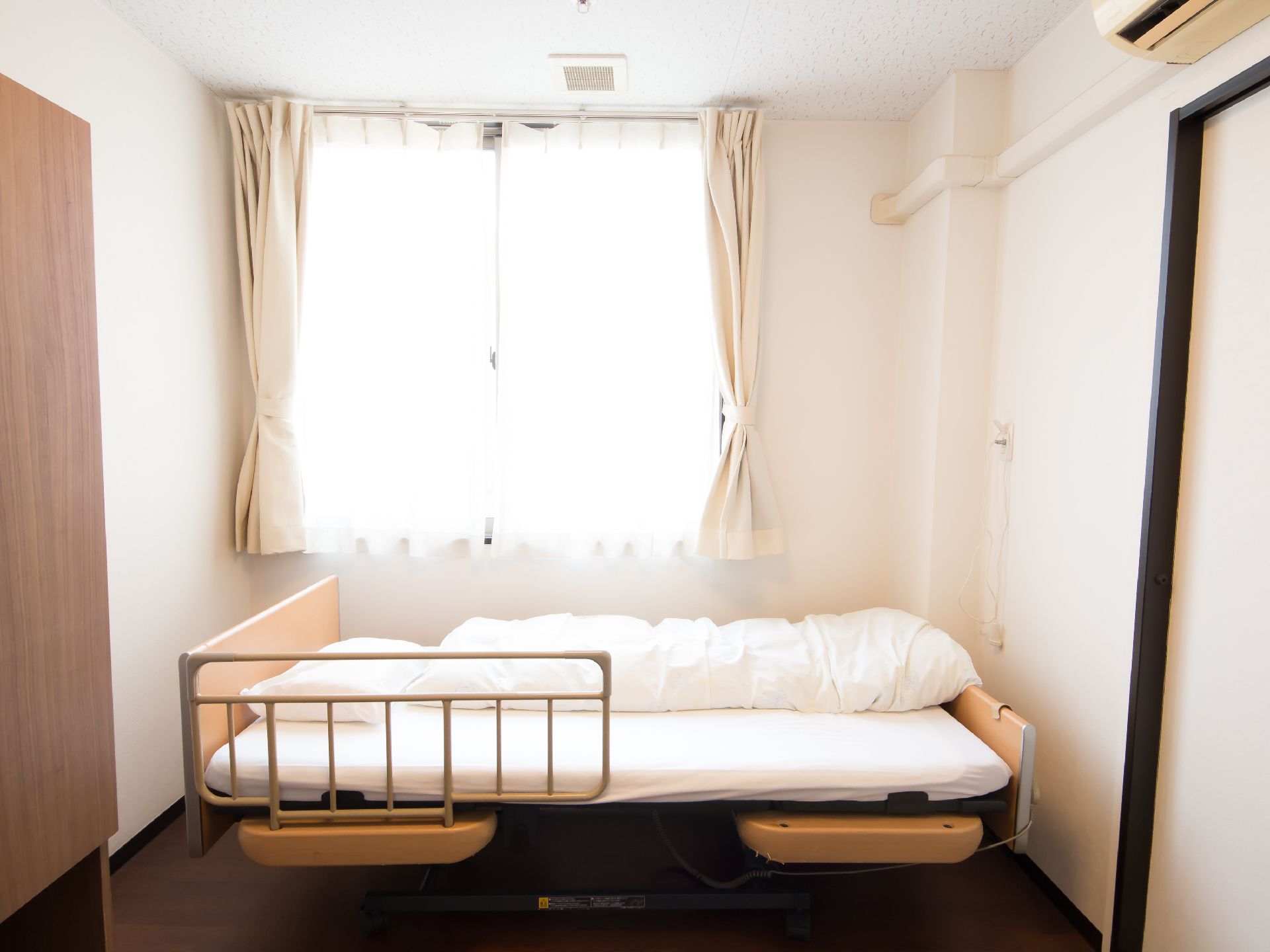 居室には大きな窓があり、ベージュのカーテンが掛けられている。窓の前に落下防止柵がついた介護用ベッドが置かれている。