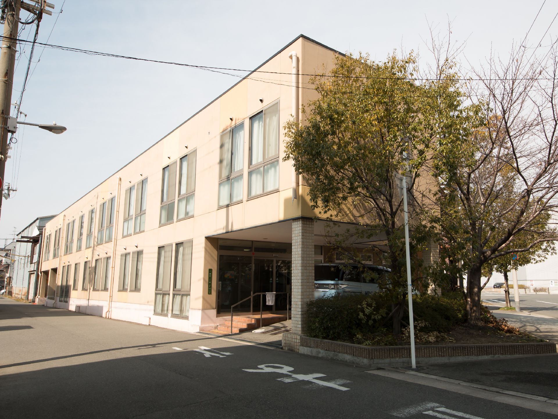 ベージュの外壁の２階建ての建物である。道路側に面した壁に居室の窓が並んでいる。建物横に大きな木が生えている。