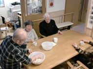 木製のダイニングテーブルで何かを作っている男性入居者とそれを見ているほかの入居者。
