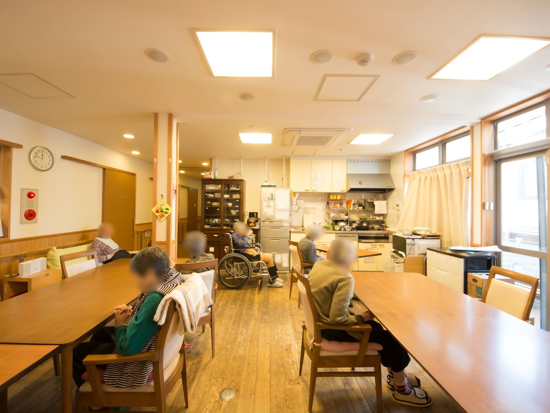 多くのテーブルや椅子が置かれた食堂がある。入居者様が６名集まって、椅子やソファ、車椅子に座っている。
