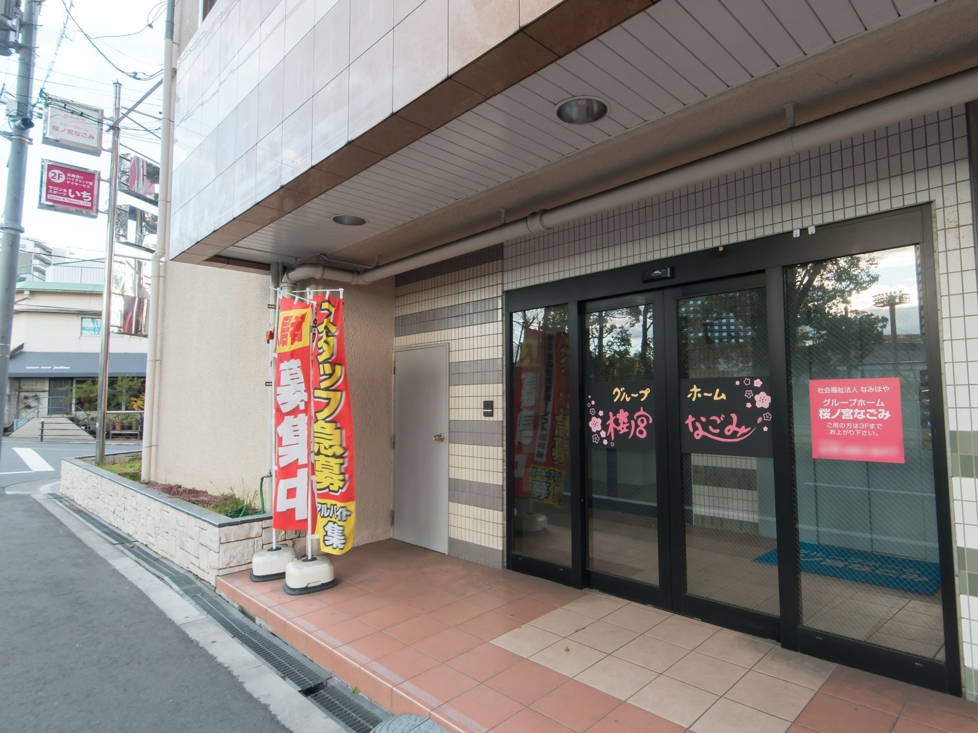 落ち着いた色合いの建物で、玄関上部の屋根部分には照明が設置されている。自動ドアの前には、旗が置かれている。