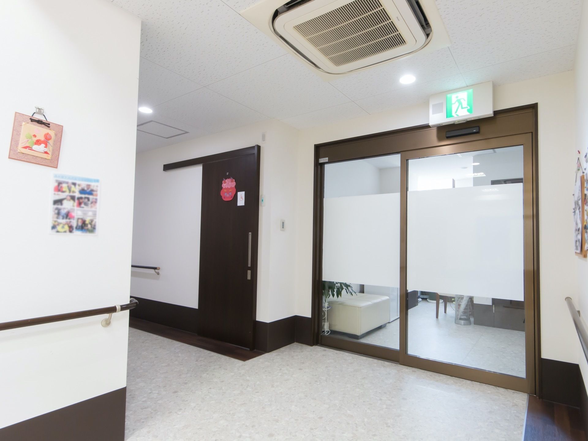 白を基調とした明るい廊下で、壁には手すりが設置されている。ガラス張りの自動ドア越しに、椅子や観葉植物が置かれているのが見える。