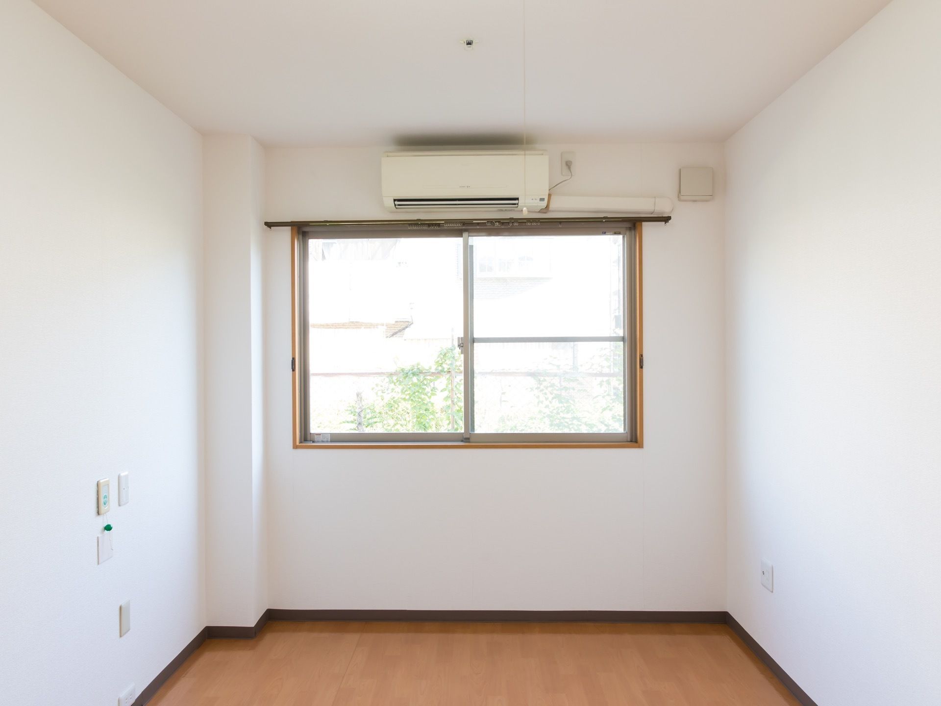 白い壁とフローリングの床の居室がある。大きな窓があり、外の景色が見える。壁にはエアコンがある。